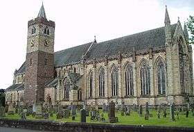 Side view of the nave of a cathedral from outside. Tall arched glass windows run along half the length of the nave from the right. Adjacent to the nave, and to the left of the scene is a cuboid-shaped tower with a conical spire. The foreground is scattered with headstones of a graveyard on green grass.