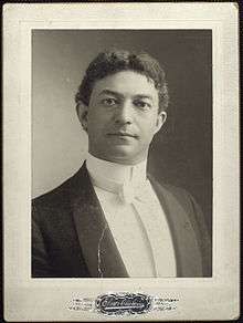 Black and white portrait of a white man with dark hair. He is wearing a tuxedo with a white bow tie.