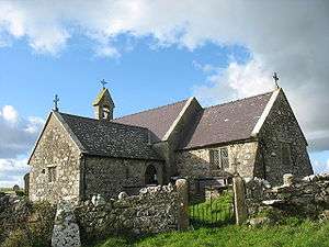 A stone church with slate roofs seen from the southeast. To the right is the chancel, to the left is a large transept, beyond which can be seen a bellcote