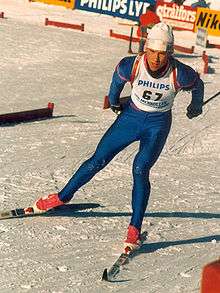 A man skies on a snow-covered course. He wears a white vest on top of a blue unitard, a white winter cap, and red ski boots. On his back, he carries a rifle.