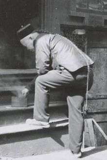 A black-and-white photograph from three-quarter back view of a man wearing a round cap and a long braided queue that reaches to the back of his right knee. His left foot is posed on the first step of a four-step wooden staircase. Bending forward to touch a cylindrical container from which smoke is rising, ahe is resting his left elbow on his folded left knee.