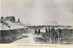 Nine men in dark clothing stand or sit around a small wooden boat that has been dragged on to a rocky shore, with rocks and icy peaks in the background.