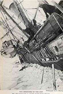  Side of a wooden steamship held in solid ice, leaning heavily to the left with a lfeboat swinging in its davits. One man visible on the ice, another aboard the ship, looking down.