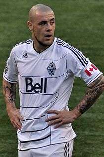 A man wearing a white t-shirt and white shorts, standing on a soccer field with his hands at on his hips, looking to his left.