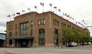 A large, rectangular, tan brick building with green trim and a roof lined with flags.