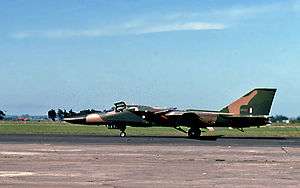 Colour photo of a jet aircraft painted in a camouflage pattern parked on concrete