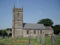 Gray stone building with square tower at left hand end. Grass and gravestones in the foreground.