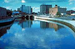 The Flint River in Flint, Michigan, United States, in the late 1970s during a U.S. Army Corps of Engineers flood control project, Taken from approximately halfway between the Grand Traverse Street bridge and Beach-Garland Street bridge, looking east. To the right in the distance is the former IMA Auditorium, later part of AutoWorld, demolished in 1997 to make way for an expansion of UM-Flint. The Northbank Center, to the right of the upper center of the photograph, is also now part of UM-Flint.