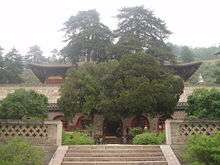 A Chinese temple building. It is built on a raised terrace and two trees nearly obscure the building from view.