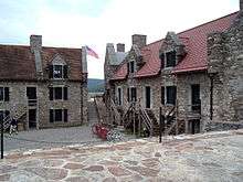 A photograph of two stone buildings with red roofs, surrounding a stone-paved central area. The buildings have entrances on two levels, with wooden stairs outside leading to the doorways on the upper level. An American flag is visible waving in the gap between the buildings.