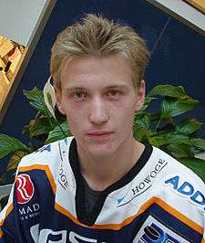 A frontal shot of an ice hockey player's head and shoulders. He has light brown hair and is wearing a blue and white uniform.