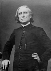 A middle-aged man with grey hair, wearing a black suit, rests one hand on the back of a chair.