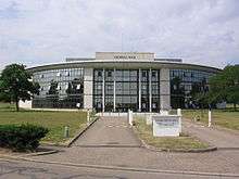 A white concrete and glass, curved-front building with a driveway separated by a median leading to it