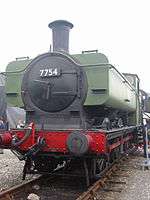 A pannier tank locomotive is seen from the front a slightly to the right. The cab and pannier tank are painted green. The chimney, smokebox, running plate, splashers, and wheels are black, and the buffer beam and coupling rods are red. The number, 7754, is shown in white letters on a black plate on the smokebox door. Part of round cab window is visible and is edged in brass. The white lettering "NCB 7754" is on the side of the pannier tank.