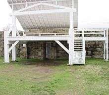 Reconstructed gallows, painted white with an open with an angled roof and brick wall at the rear.