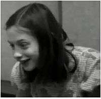 A black and white frame taken from a video of Genie, who is enthusiastically smiling. It shows her from the chest up, taken from several feet away while Genie is facing slightly to the right of the camera.