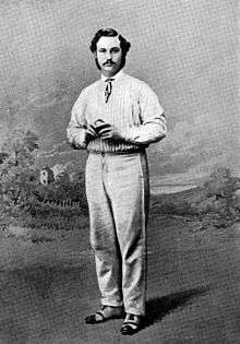 A cricketer holding a ball posing before a painted backdrop of a field