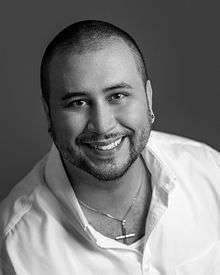 Black and white portrait of George Zimmerman smiling with an open white collared shirt and gold cross chain