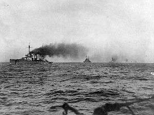 A line of large warships. Thick black smoke pours from their funnels as they steam through choppy seas.