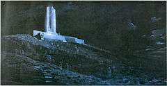 A crowd of dark and ghostly soldiers are scattered on a hill of churned ground, shell holes, and general battlefield detritus. A memorial, painted in white, is located at the top of the hill.