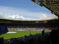 A stadium full of spectators. Those nearest the camera are waving blue and white flags.