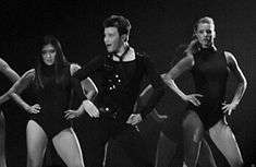 The grayscale picture of three people, two women and a man, who dance and put their hands on their hips. The man wears a dark outfit, compound of a vest, a shirt and pants. The women wear similar leotards.