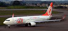 Mid-sized twin-engine passenger jet with red-and-white GOL logo, whose wings end in 2-meter upturned winglets, on an airport taxiway. A lineman is crouching next to the nose gear.