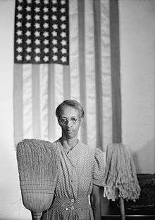 Man holding up a broom and mop with an American flag hanging in the background, in imitation of the original American gothic.