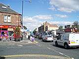 The junction of High Street and Church Road. A terrace of stone fronted buildings, with a curved corner building fronted in white, from the mid-late 19th century. The modern shop fronts are not in keeping the original style of the buildings. It is set back from the road by modern paving. The far section of the building is the old fire station building (formerly Urban District offices).