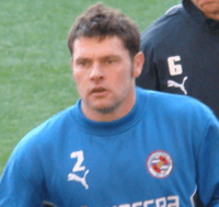 A man with short brown hair is wearing a blue top. He is standing on a grass field.