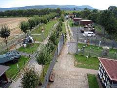 Elevated view of a series of huts and vehicles exhibited in the open air on either side of a stretch of concrete patrol road, with a range of forested hills visible in the background.