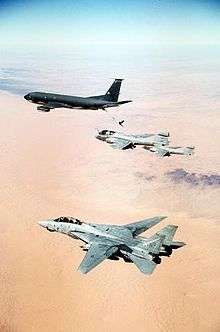 Portrait photography of four aircraft overflying orange desert and almost-flat terrain; horizon is blurred. Leading is black aircraft, followed by two single-engine jet aircraft, the one closer to camera being refueled by leading jet via a stiff hose connecting the two. Closest jet to camera is pale gray, has two engines and vertical fins, flying with wings unswept.