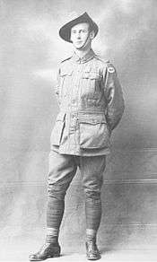 A full body portrait of a standing man. He is wearing a military uniform, a slouch hat and has a medal pinned to his left breast. His arms are behind his back and his right leg is forward. Three stripes can be seen on his right sleeve, denoting his rank as sergeant.