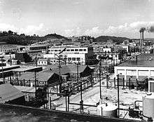 An array of industrial buildings with lots of power poles and wires. Are a pair of smokestacks is in the background.