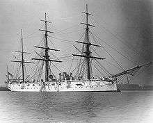 Starboard broadside view of a metal sailing ship in port at anchor. The three masts are rigged with yards and furled sails. A smokestack is amidships, and ventilators rise above the top deck. Guns are visible protruding through the sides of the ship.