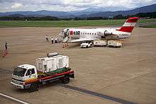 A Bombardier CRJ-200 aircraft parked on the tarmac with passengers approaching the aircraft for boarding. There is also a baggage trolley and two carts next to the aircraft. There is an auxiliary power unit in the foreground and a mountain view in the background