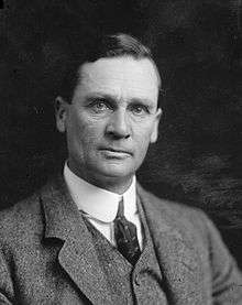 Shows a clean shaven man looking towards the camera. He wears a shirt and tie, a waistcoat, and a suit jacket.