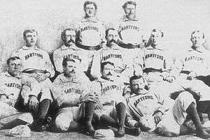 In this photograph of a baseball team, eleven men are situated in three rows facing the camera, with four sitting on the floor, five sitting in chairs, and two standing.