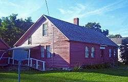 A 1 1⁄2-story wooden house, painted red, lit by the sun from the left with trees and part of a white house behind it.