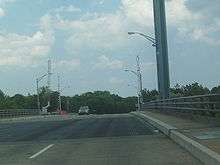 A long bridge with a steel grate deck and lift towers gazing over cars and a pedestrian, who is on the side of the bridge jogging