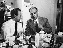 A young man in a white shirt and tie and an older man in suit and tie sit at a table, on which there is a tea pot, plates, cups and saucers and beer bottles.