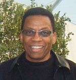 An African American male in his late sixties is wearing sunglasses, a black jacket, and a black T-shirt.