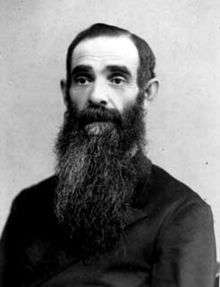 A man with a long dark beard and a dark shirt sits and looks straight ahead