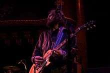 Huw Bunford playing guitar while looking to his right, away from the camera. The guitarist has a large beard and is wearing a brown jacket.