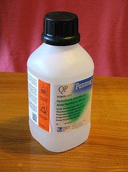 White plastic bottle with safety cap is labeled "QP Panreac" above smaller text "Hydrofluoric Acid 40% QP" with 6 translations. In a bright orange region along the side, warning symbols are clearly visible.