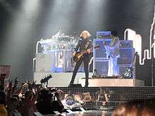 A female blond performer playing an electric guitar on stage. She is wearing black jacket and pants. The backdrops behind her display a number of tall buildings. The stage is illuminated by a number of halogen lights from the ceiling.