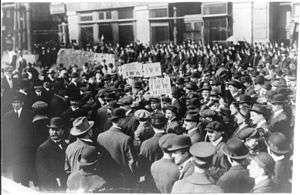 Black and white photograph of a large crowd of people, a few holding signs above the crowd, displaying IWW acronyms and slogans.