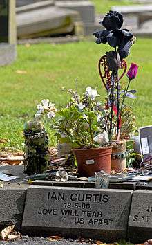 A grayish stone block with "Ian Curtis 18-5-80 Love Will Tear Us Apart" carved into it in a sans-serif typeface. There are several small pots of flowers and other objects on top.