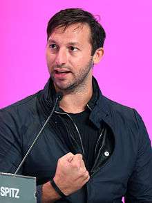 Young man with short blonde-brown hair wearing a dark open necked shirt and zip-up jacket, not cleanly shaven, smiling broadly. The background is labeled with the word 'Brasserie'.