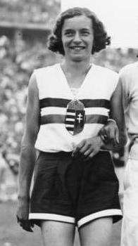 A black and white photograph of a female athlete with short cut hair. She wears a white sleeveless top with two horizontal stripes and a crest in the middle of her chest, and dark shorts.